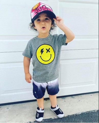 Toddler boy wearing a grey smiley face shirt and blue and white shorts in the driveway.