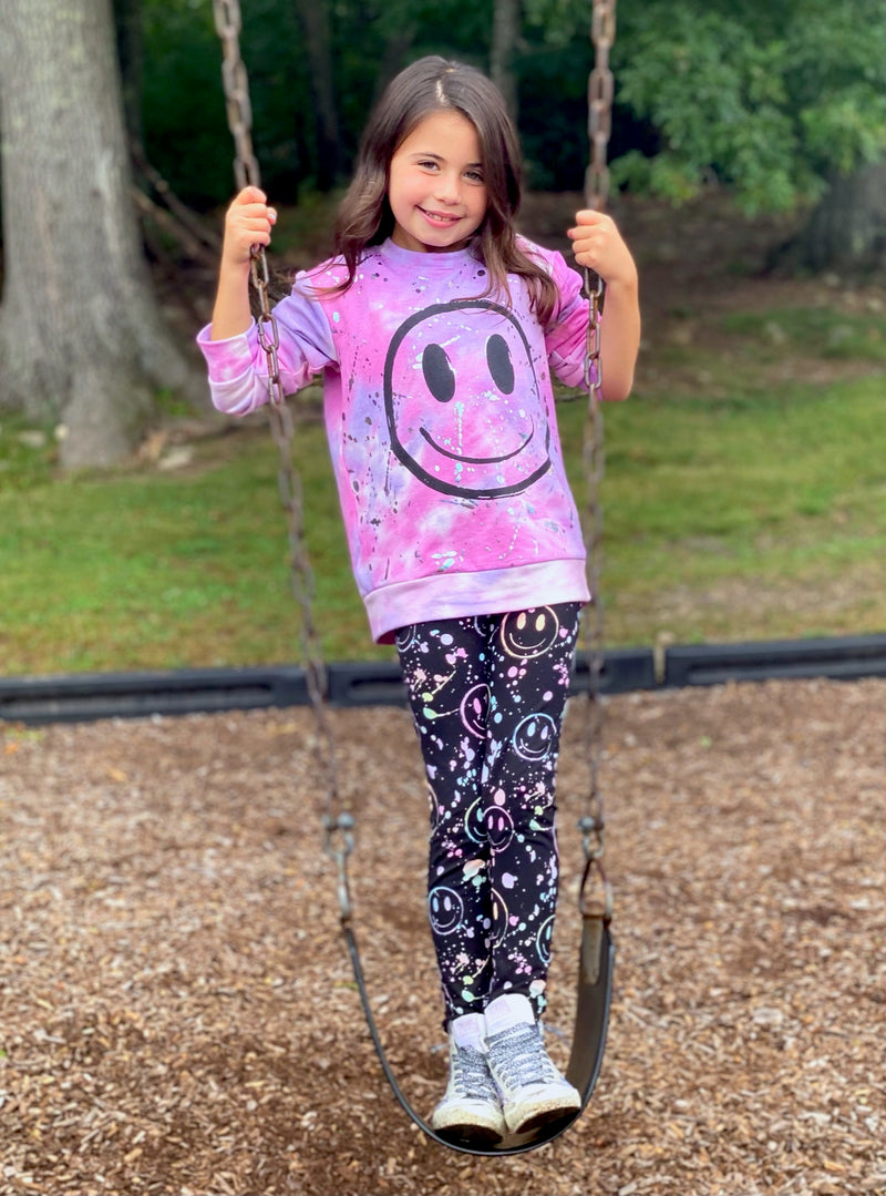 Girl standing on the sitting portion of a swing set wearing a pink smile face shirt.