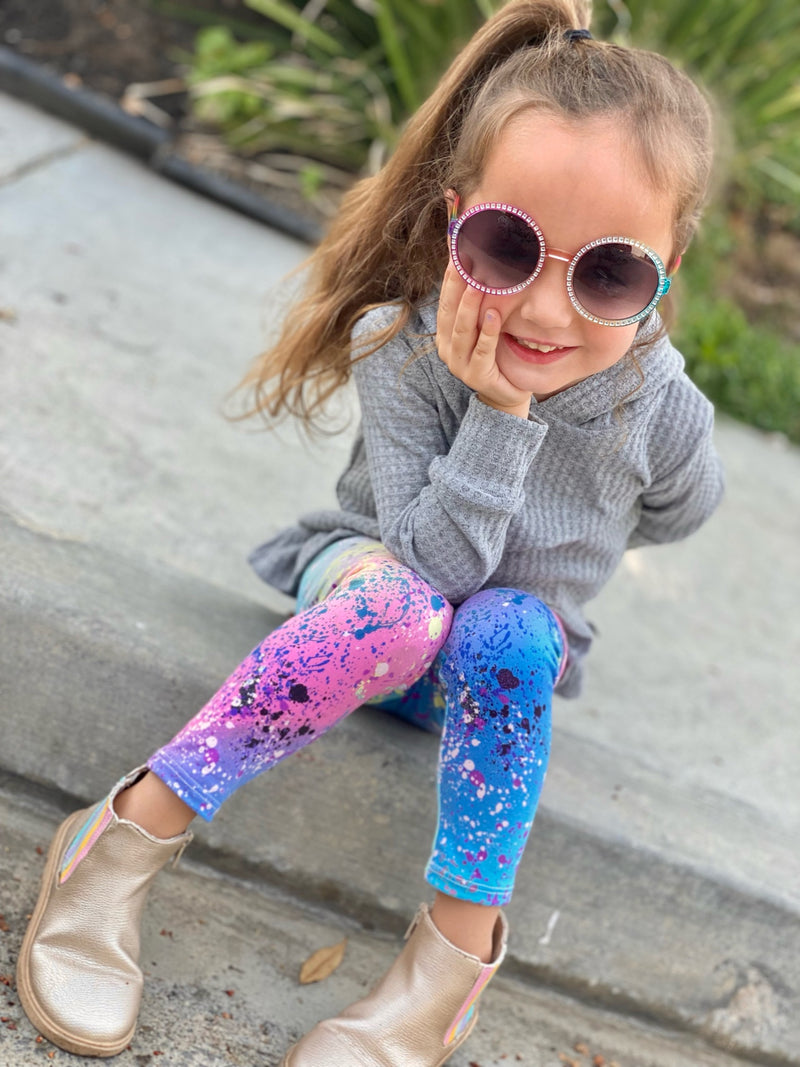 Young girl wearing sunglasses and pink and blue leggings while sitting on the curb.